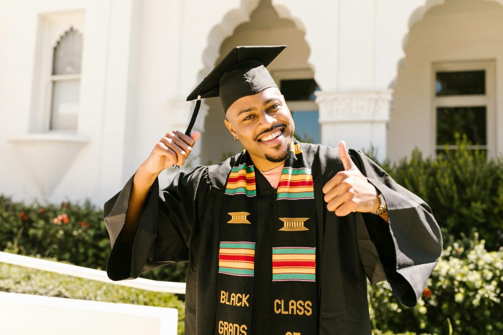 Bristol University Scholarships. Smiling graduate in cap and gown with kente stole giving thumbs up at outdoor ceremony.